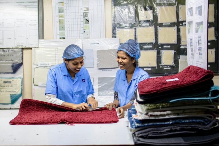 Home Two female textile workers inspecting fabrics with cheerful expressions in a factory setting.